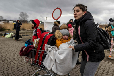 Aufnahme Ukrainerinnen auf der Flucht, Frau mit Kleinkind in Einkaufswagen, vollgepackt mit Koffern und anderen Habseligkeiten, dahinter weitere Menschen auf der Flucht