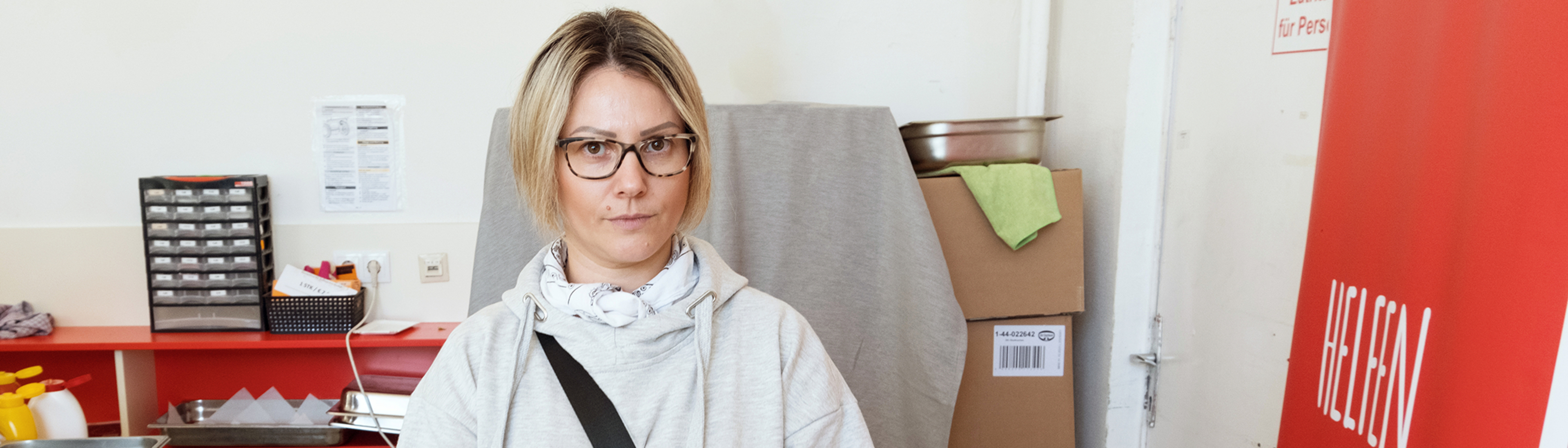Frau im grauen Kapuzenpulli bei Samariter Suppentopf, kurze Blonde Haare, Brille, hält Teller mit Apfelstrudel, daneben rotes Roll up 