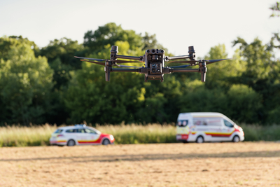 Aufnahme einer Drohne, die über einem Feld fliegt, dahinter unscharf zu erkennen zwei Fahrzeuge des Samariterbund Wiens, im Hintergrund Wald mit Bäumen