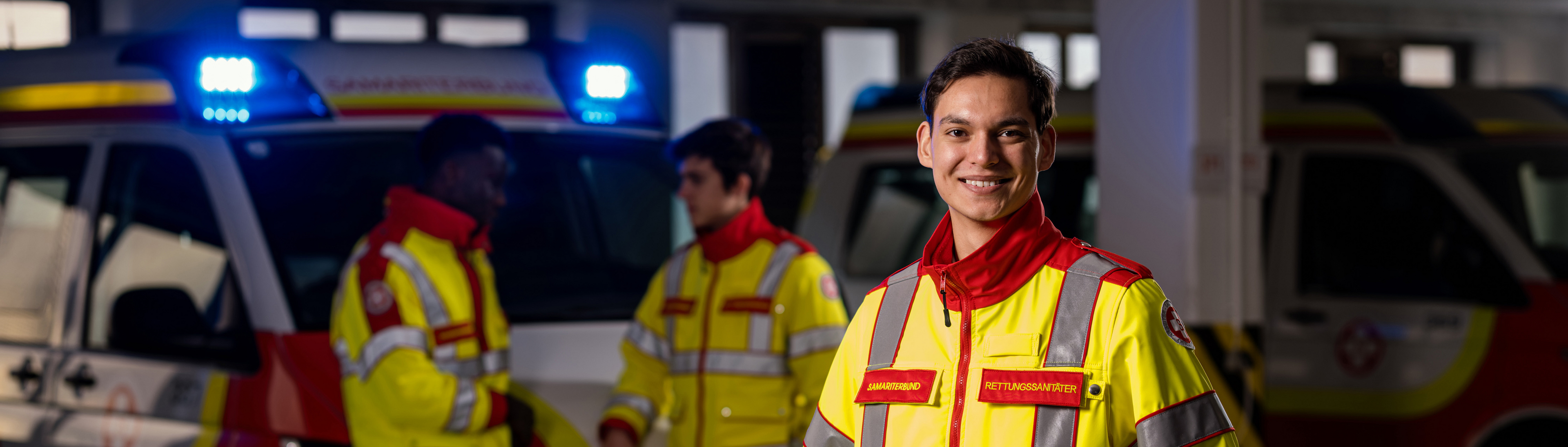 Im Vordergrund steht ein lächelnder Zivildiener in Uniform, während zwei weitere Zivildiener vor dem Rettungswagen im Hintergrund stehen. Das eingeschaltete Blaulicht des Rettungswagens sorgt für eine coole Atmosphäre.