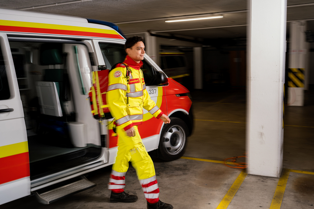 Der Zivildiener in Uniform trägt seinen Einsatzrucksack zum Einsatzort. Im Hintergrund ist ein Rettungswagen mit eingeschaltetem Blaulicht zu sehen.