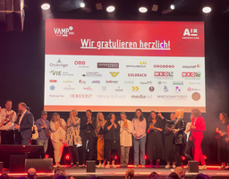 Foto aufgenommen bei Preisverleihung VAMP Award, Blick auf Bühne, viele Menschen stehen dort und freuen sich, klatschen in die Hände, im Hintergrund Leinwand mit Spruch Wir gratulieren herzlich und viele Sponsorenlogos 