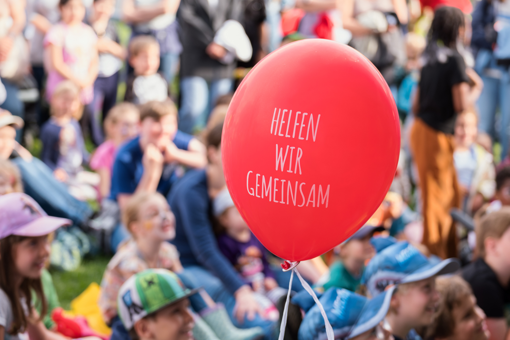 im Fokus roter Luftballon mit Spruch Helfen wir gemeinsam, im Hintergrund verschwommen Kinder vor Bühne