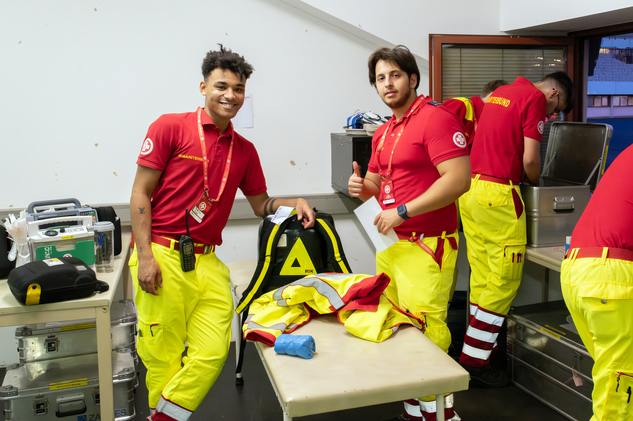 Zwei Rettungskräfte in roten Shirts und gelben Hosen lachen in einem Einsatzraum.