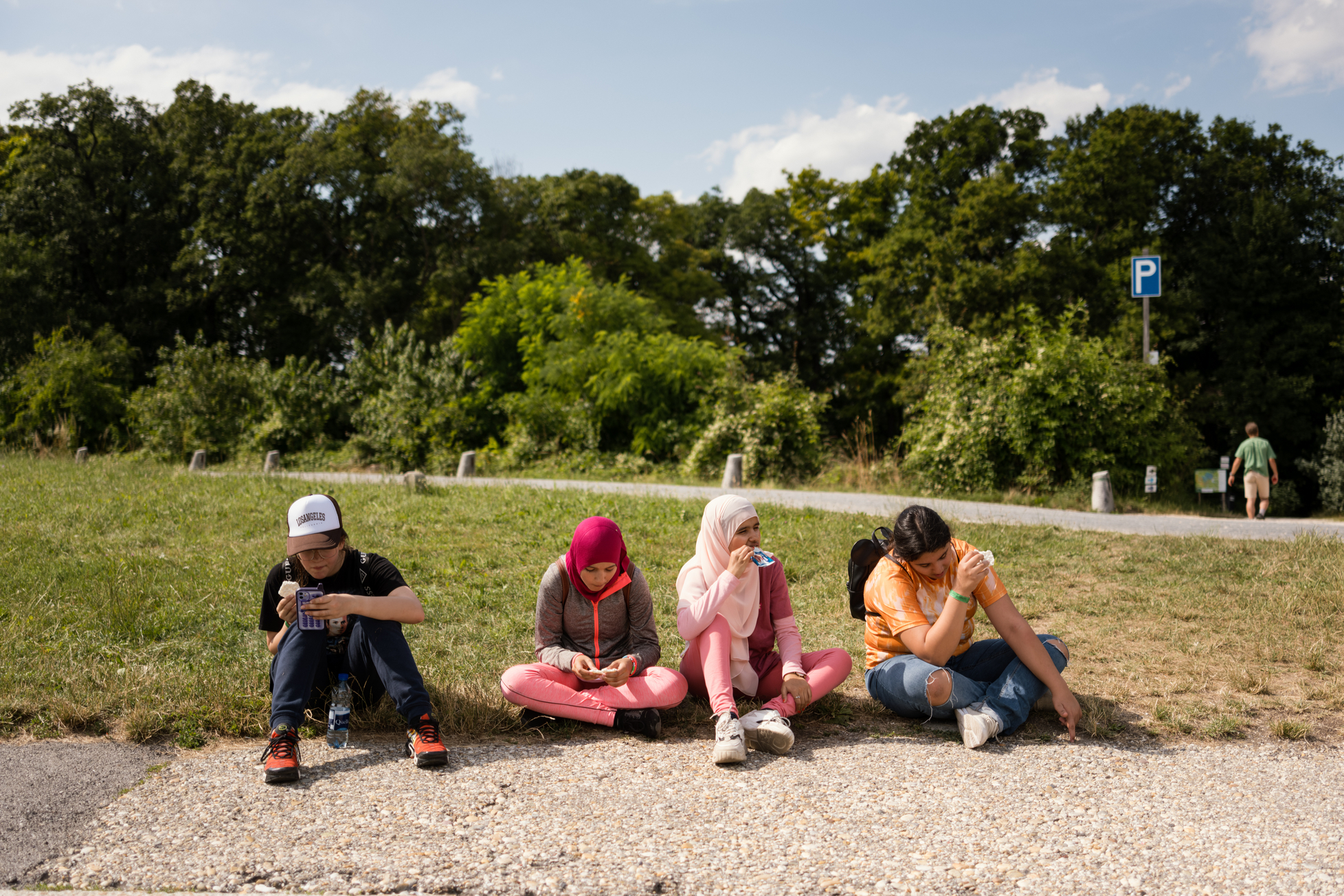 vier Kinder sitzen auf Rasen, davor Schotterweg und machen eine Pause und essen eine Jause