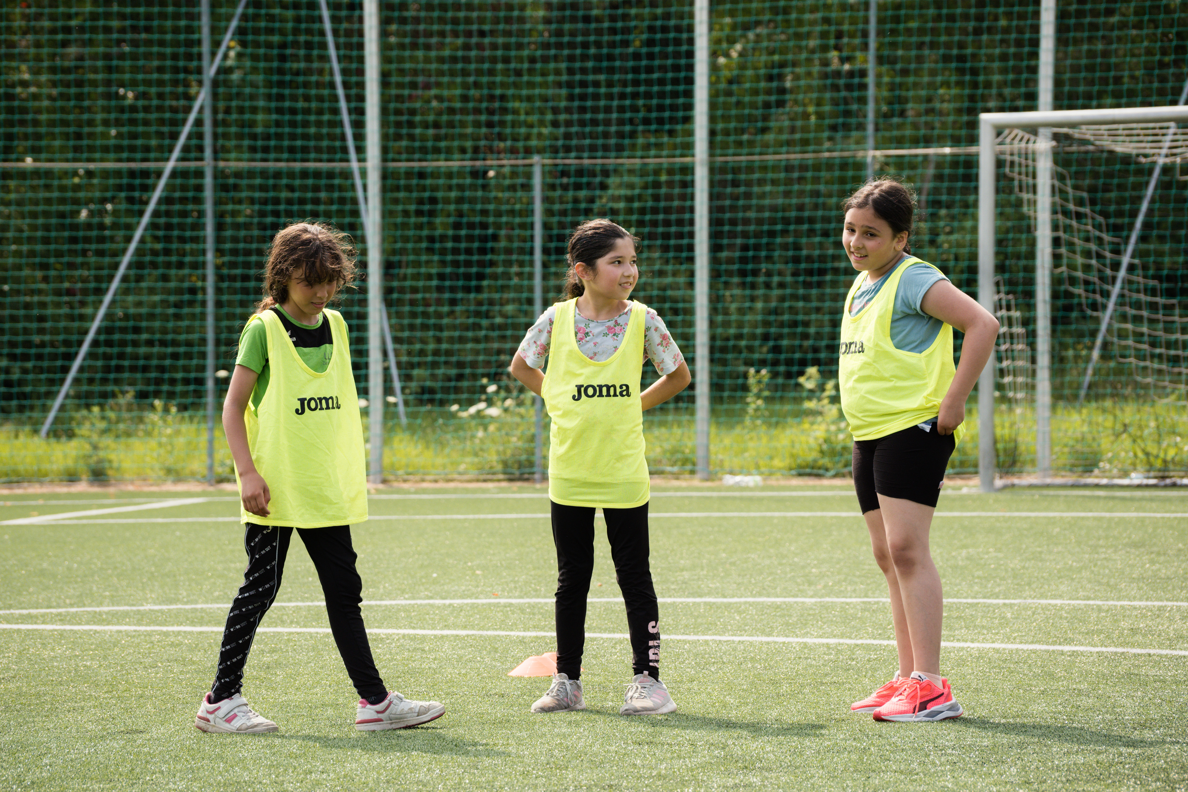 drei Mädchen mit gelben Trikots auf einem Fußballfeld, schauen sich an und reden miteinander, rechts hinten Tor zu erkennen