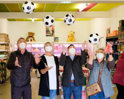 mehrere Personen in Sozialmarkt, alle tragen FFP2-Maske und werfen Fußbälle in die Luft, im Hintergrund Supermarktatmosphäre mit Kühlregal und weiteren Lebensmittelregalen