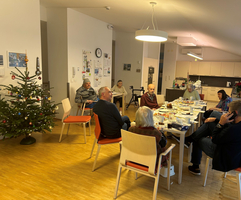 mehrere Personen in einem Raum, sitzen rund um Tisch auf Sessel, Senioren WG, Tisch ist gedeckt, links sieht man einen Christbaum, der dekoriert ist