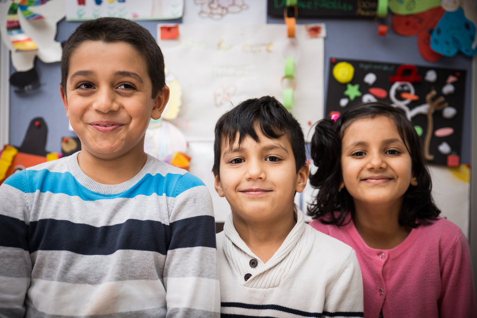 Drei lächelnde Kinder stehen vor bunten Bastelarbeiten.