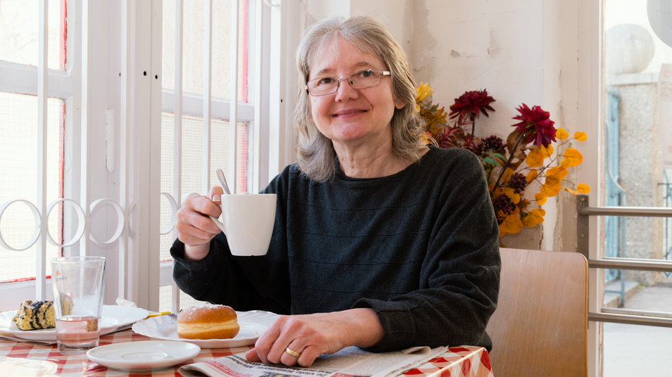 Eine lächelnde Frau mit grauen Haaren sitzt an einem Tisch mit rot-weißem Tischdecken. Sie hält eine Tasse in der Hand und hat einen Donut und ein Glas Wasser auf dem Tisch. Im Hintergrund sind dekorative Blumen zu sehen.