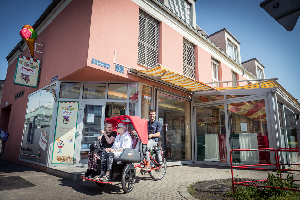Einen Mann, der eine Fahrrad-Rikscha mit zwei Fahrgästen vor einem Geschäft fährt.