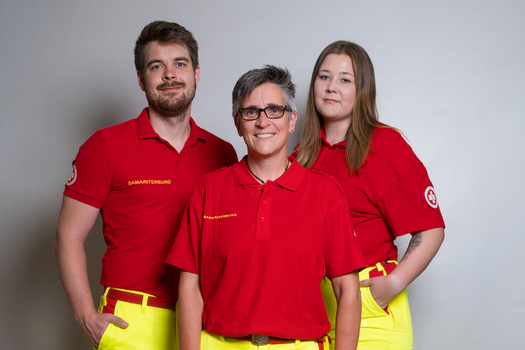 Drei Personen in roter Oberbekleidung und leuchtend gelben Hosen stehen zusammen.