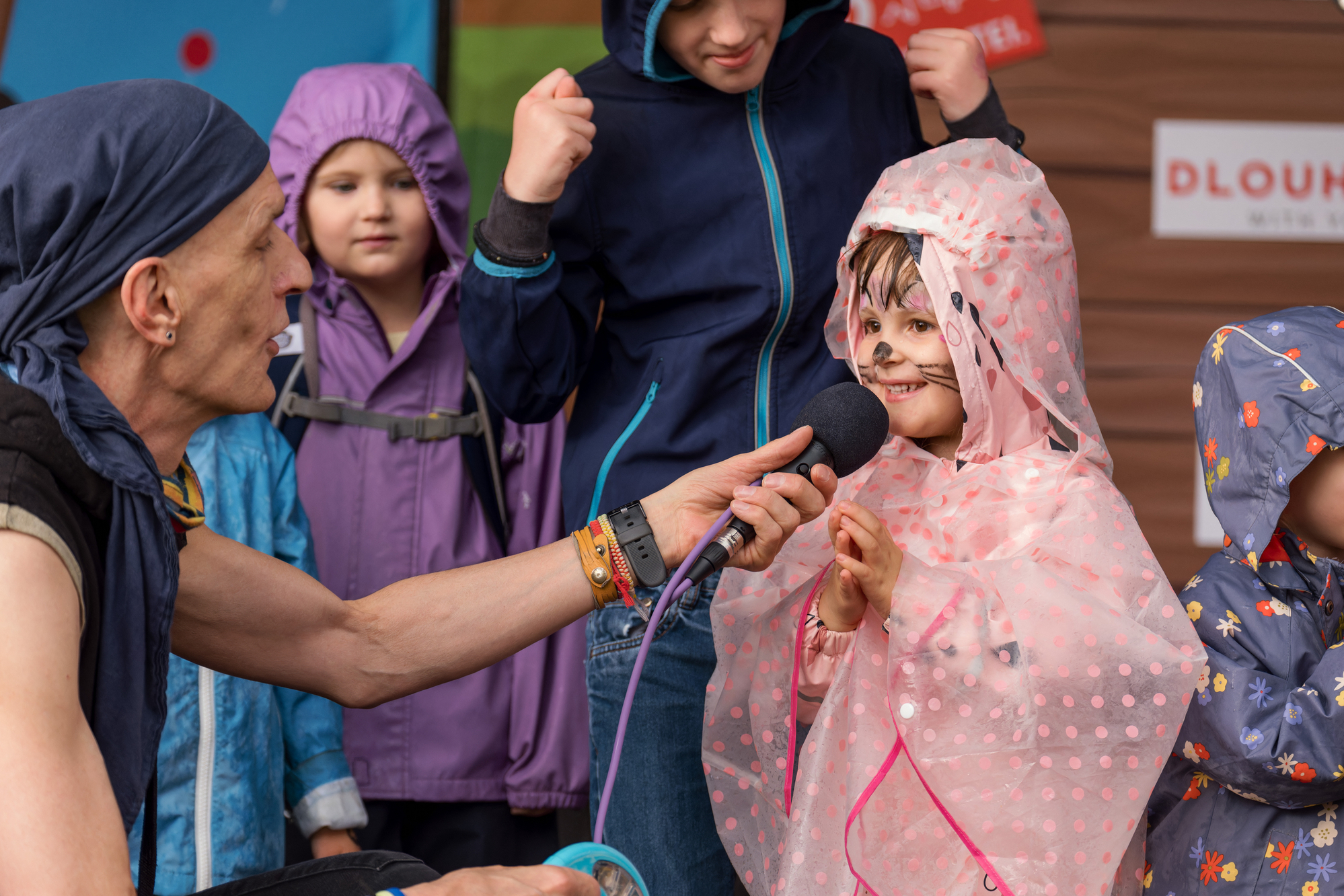 Kinderliedermacher Bernhard Fibich auf Bühne am Tag des Samariterbundes, hält Mikrofon in der Hand, Kind mit Katzenbemalung und Regencap, dahinter weitere Kinder