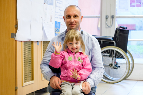 Vater im Rollstuhl hat Tochter am Schoß und sie winkt in die Kamera