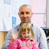 Vater im Rollstuhl und Tochter auf seinem Schoß winkt in Kamera