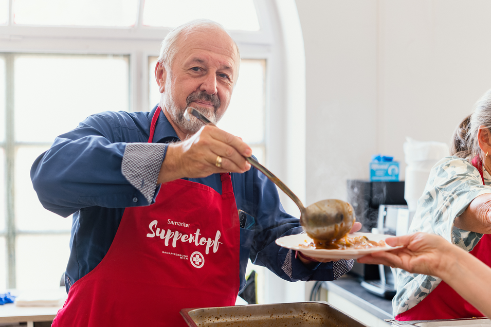 Ein Mann mit einer roten Schürze steht in einer Küche und lächelt in die Kamera. Er hält einen großen Schöpflöffel in der Hand und serviert eine herzhafte Suppe oder einen Eintopf aus einem großen Behälter.