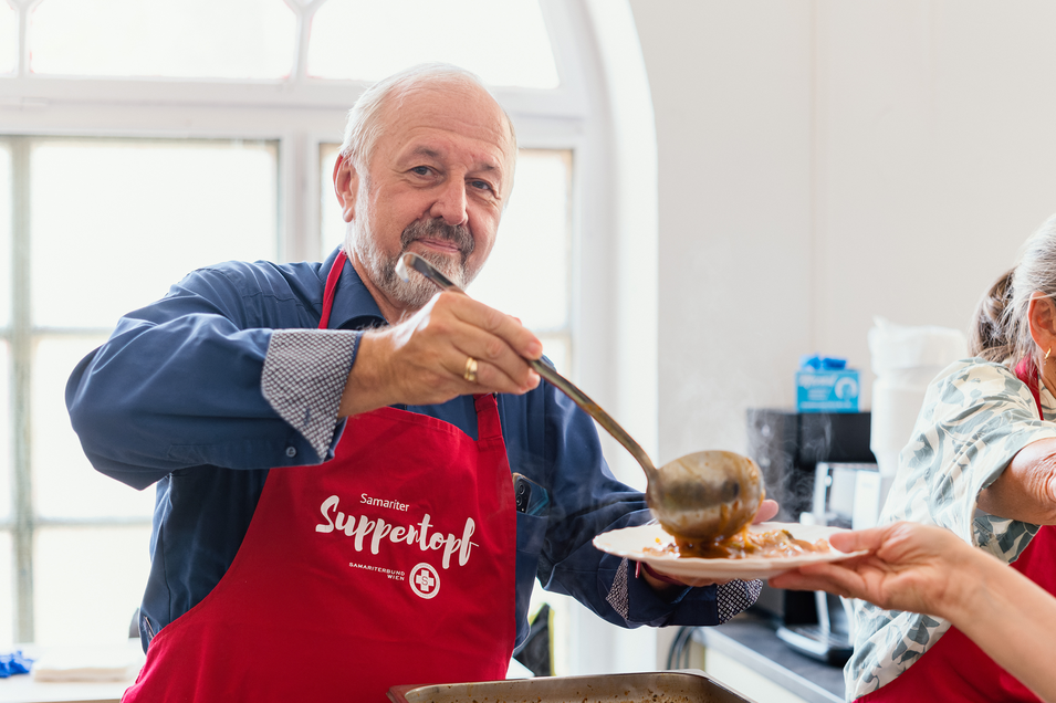 Ein Mann mit einer roten Schürze steht in einer Küche und lächelt in die Kamera. Er hält einen großen Schöpflöffel in der Hand und serviert eine herzhafte Suppe oder einen Eintopf aus einem großen Behälter.