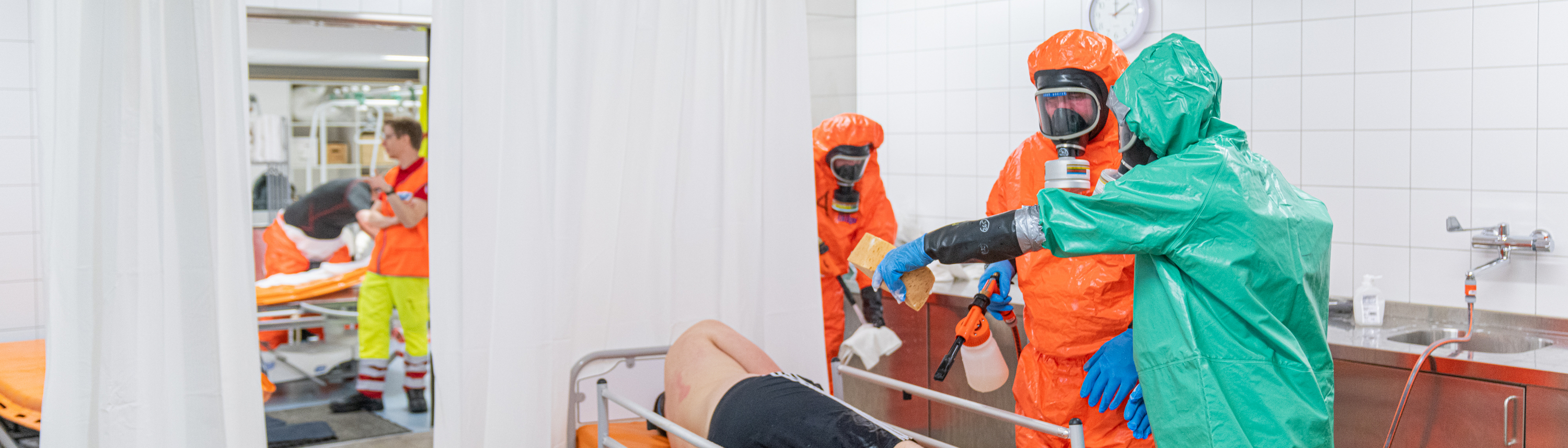 Ein Patient liegt im Zuge des Katastrophenschutzes in einem Zelt, Mitarbeiter:innen in Schutzanzügen umsorgen ihn.