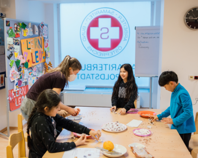 Aufnahme in LernLEO, Kinder die an einem Tisch Kekse backen, Betreuerin unterstützt sie, im Hintergrund Pinnwand mit Zeichnungen, Glaswand mit spiegelverkehrtem Samariterbund Logo