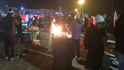 Eine Gruppe von Menschen steht in der Nacht bei einer Tonne in der ein Feuer brennt. Im Hintergrund steht ein voll besetzter Bus.