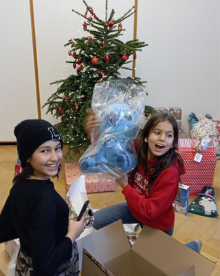 zwei LernLeo Mädchen packen gerade Geschenke vor einem Christbaum aus und lächeln dabei in die Kamera.