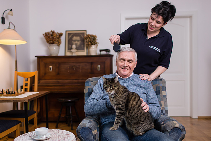Heilhelferin bürstet älterem Herrn in seinem Wohnzimmer die Haare und er streichelt Katze auf seinem Schoß
