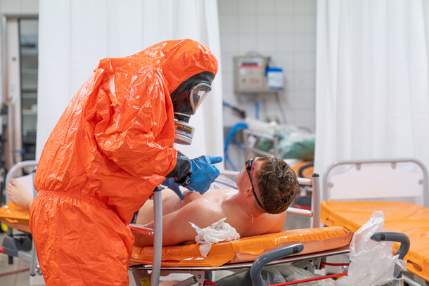 Mann liegt auf einer Trage, er trägt eine Schwimmbrille. Zweiter Mann hat orangenen Schutzanzug an mit Maske und Handschuhe.