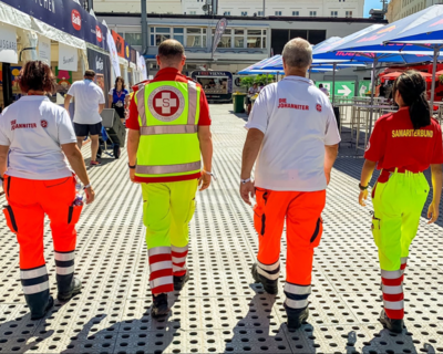 vier Sanitäter:innen von hinten fotografiert, links außen und zweite von rechts Sanitäter:innen der Johanniter, rechts außen und zweiter von links Sanitäter:innen von Samariterbund Wien - alle vier in Uniform, Logo jeweils am Rücken erkennbar