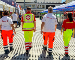 vier Sanitäter:innen von hinten fotografiert, links außen und zweite von rechts Sanitäter:innen der Johanniter, rechts außen und zweiter von links Sanitäter:innen von Samariterbund Wien - alle vier in Uniform, Logo jeweils am Rücken erkennbar