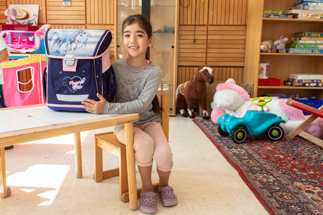 Schulanfängerin sitzt im Gemeinschaftsbereich ihrer Flüchtlingseinrichtung und freut sich über ihre neue Schultasche