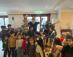 Ein Mann mit Weihnachtsmannmütze steht vor einem Christbaum vor ihm stehen viele Kinder.