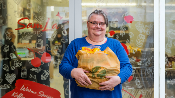 Eine Sozialmarktkundin steht mit einem gefüllten Einkaufssackerl vor dem Sozialmarkt und Lächelt