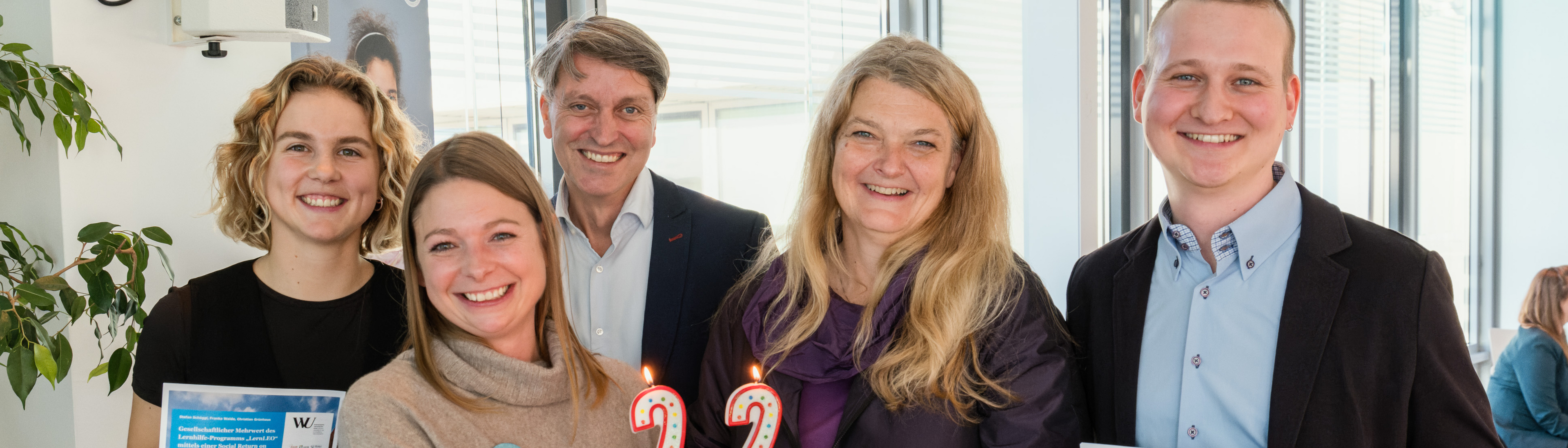 Auf dem Bild sind 3 Frauen und 2 Männer zu sehen. Zwei Personen in der Mitte halten eine Torte, die mit Kerzen verziert sind, die die Zahl 22 darstellen. Zwei Personen am Rand halten einen Folder von einer Studie der WU.