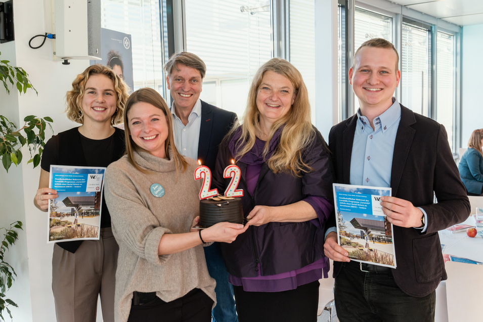 Auf dem Bild sind 3 Frauen und 2 Männer zu sehen. Zwei Personen in der Mitte halten eine Torte, die mit Kerzen verziert sind, die die Zahl 22 darstellen. Zwei Personen am Rand halten einen Folder von einer Studie der WU.