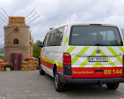 Ein Auto vom Samariterbund, dass auf einer Straße geparkt steht und das man von hinten sieht. Dahinter sieht man eine Mühle und Heubalken. 