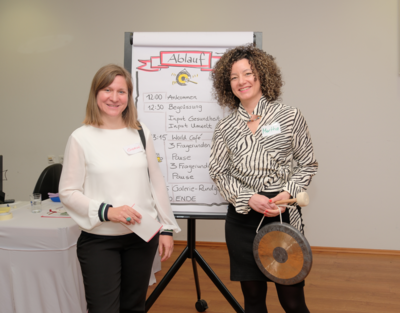 Zwei Frauen vor Flipchart, lächeln, eine hält Gong in der Hand