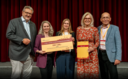 Fünf Personen stehen auf der Bühne und halten einen Gewinn in der Hand.