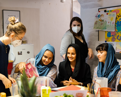 Aufnahme von Smoothie-Workshop im LernLEO im Fokus drei Mädchen, die an einem Tisch sitzen und alle lächeln, im Hintergrund bzw. daneben Betreuerinnen mit FFP2-Maske