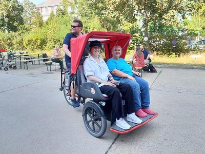 Rickscha Ausfahrt beim Sommerfest des Haus Riga, eine Rikscha auf der zwei Personen sitzen und in die Kamera lächeln, Fahrer mit Brille blickt seitlich. Dahinter Biertischgarnituren und Pflanzen