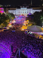 Eine große Menschenmenge versammelt sich bei Nacht vor einem erleuchteten Gebäude, umgeben von Bäumen und Ständen. Bunte Lichter erzeugen eine festliche Atmosphäre, während der Platz mit Besuchern gefüllt ist.