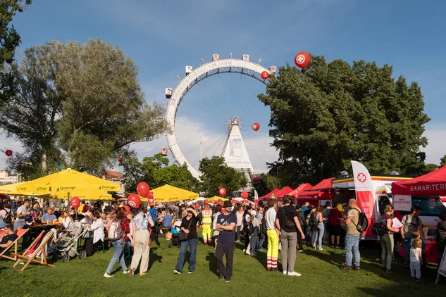 Aufnahme von Tag des Samariterbundes mit Blick auf Riesenrad, viele Menschen auf der Kaiserwiese, gelbe Ottakringer Schirme, rotes Samariterbund Zelt, im Hintergrund Bäume und blauer Himmel