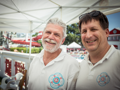 zwei Männer in weißen Polo-Shirts mit Logo der Wasserrettung stehen auf beschatteter Terrasse, im Hintergrund ein Freibad