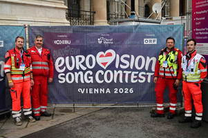 Vier Sanitäter in roten Uniformen stehen vor einem großen Banner des Eurovision Song Contest 2026 in Wien. Das Banner zeigt den Wettbewerbstitel und das Stadtwappen. Die Sanitäter lächeln in die Kamera.
