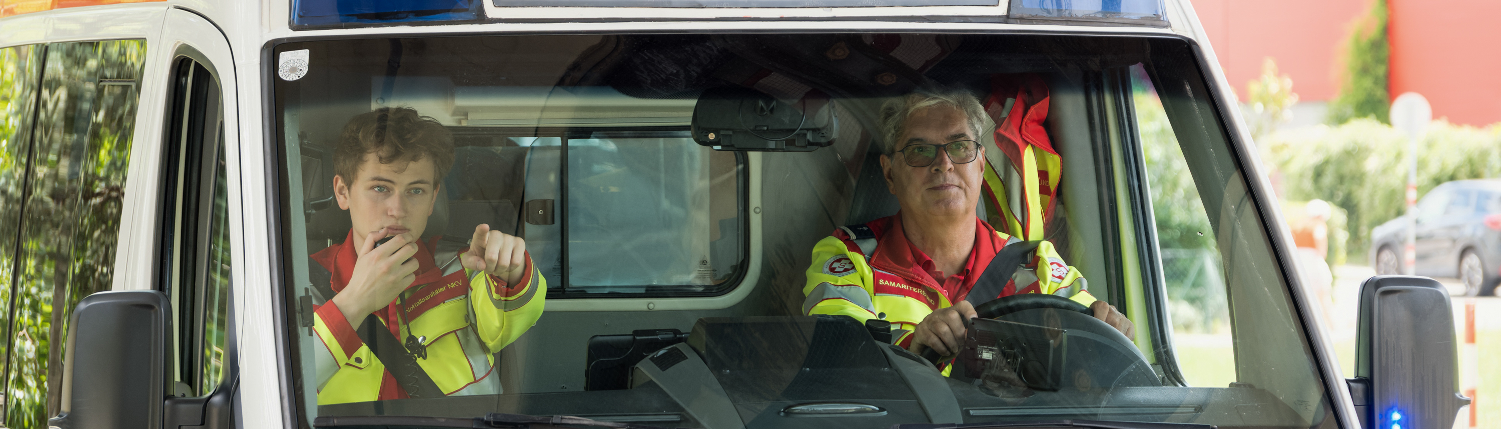 Frontansicht von Rettungswagen in dem ein junger und ein Sanitäter mittleren Alters sitzen 