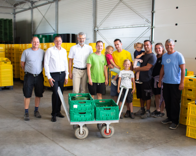 Gruppenfoto in Lagerhalle, im Vordergrund auf Rollwagen grüne Kisten mit Gurken, im Hintergrund gelbe Kisten