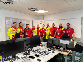 Eine Gruppe von Rettungskräften in roten und gelben Uniformen steht in einem Büro. Im Hintergrund sind mehrere Computer und Arbeitsplätze sichtbar. Die Personen lächeln und posieren zusammen, während sie Ausweise tragen.