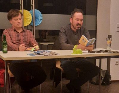 zwei Männer, links Bernd Watzka, rechts Bernd Remsing, mit Buch in der Hand, sitzen an einem Tisch vor einer Glaswand, Internetcafé, ZwischenSchritt, Lesung