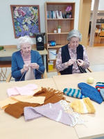Zwei ältere Frauen stricken gemeinsam bunte Schals an einem Tisch.