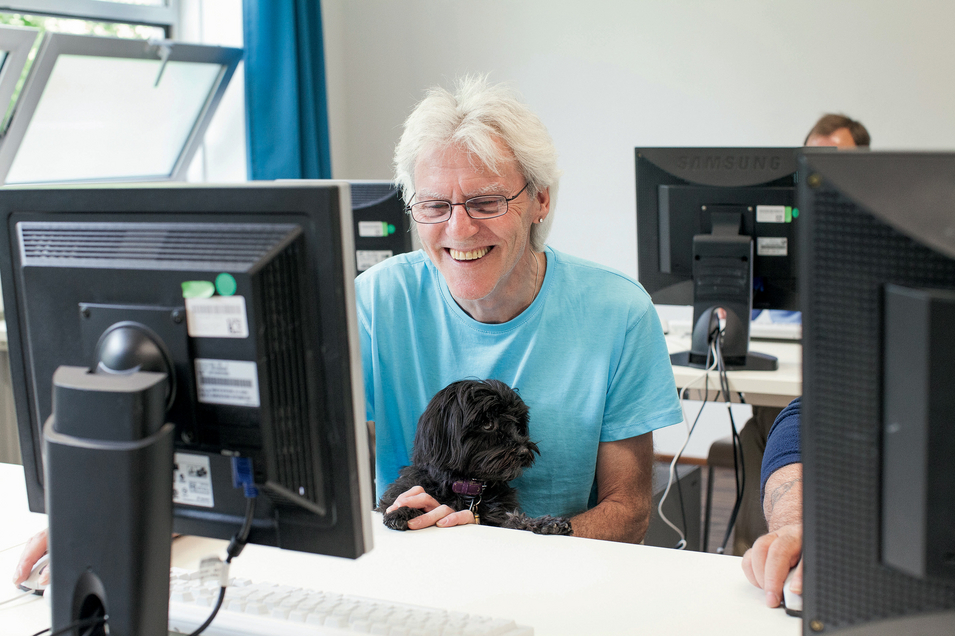 Internetcafé Zwischenschritt - Mann sitzt mit Hund am Schoß hinter Bildschirm und lächelt