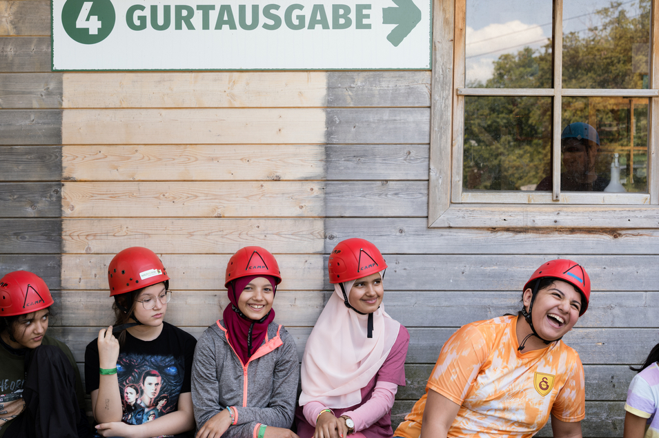 mehrere Kinder aus PowerLEO bei Hochseilklettergarten, sitzen auf einer Bank, haben Sicherheitshelme auf in der Farbe rot und lachen